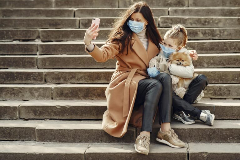 Full body of female and adorable little girl in casual clothes and medical masks sitting on steps with toy in mask and cute dog taking photo on smartphone